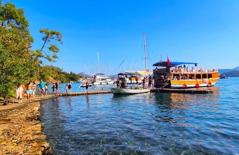 Tur teknelerinin demirlediği berrak deniz ve yürüyüş iskelesiyle huzurlu bir sahil manzarası.