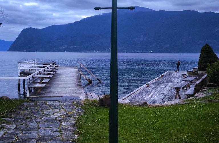 Göl kenarında çiçeklerle çevrili taş yol ve aydınlatma lambasıyla huzurlu akşam manzarası.